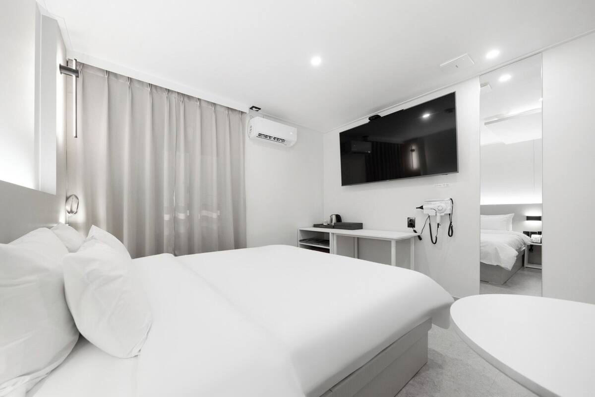 Ein modernes Hotelzimmer mit einem großen Bett, einem Flachbildfernseher an der Wand, einem Nachttisch mit Lampe sowie einem integrierten Schminktisch mit Spiegel.