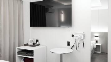 A modern hotel room with a flat-screen TV mounted above a white sink, a hairdryer on the wall, and a small bedside table with a lamp.