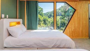 A modern bedroom with a large bed, a headboard with a light, and a view of a forest through a large window.