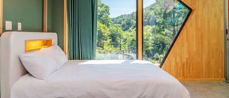A modern bedroom with a large bed, a headboard with a light, and a view of a forest through a large window.