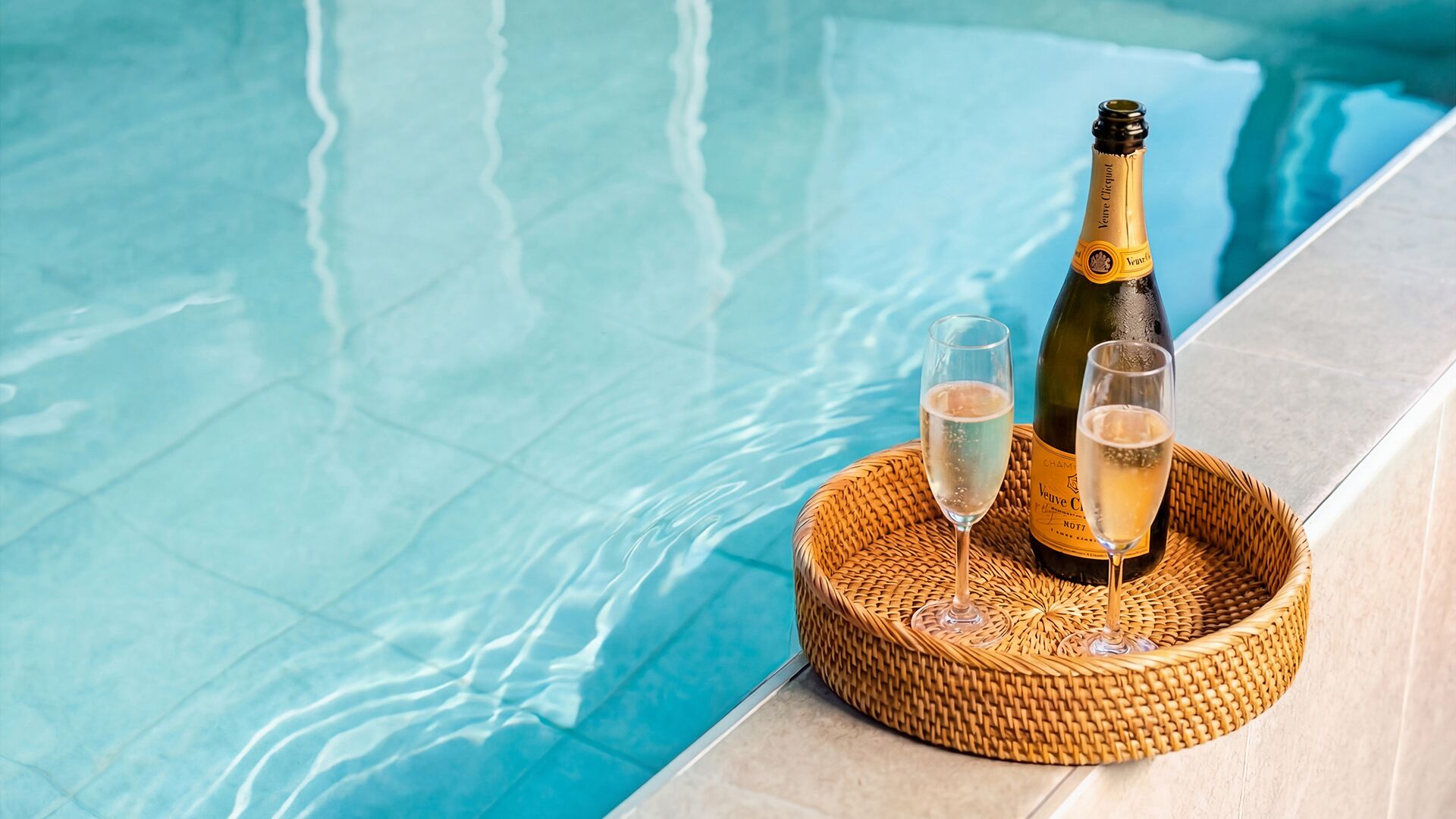 A bottle of champagne and two glasses on a woven tray by a pool.