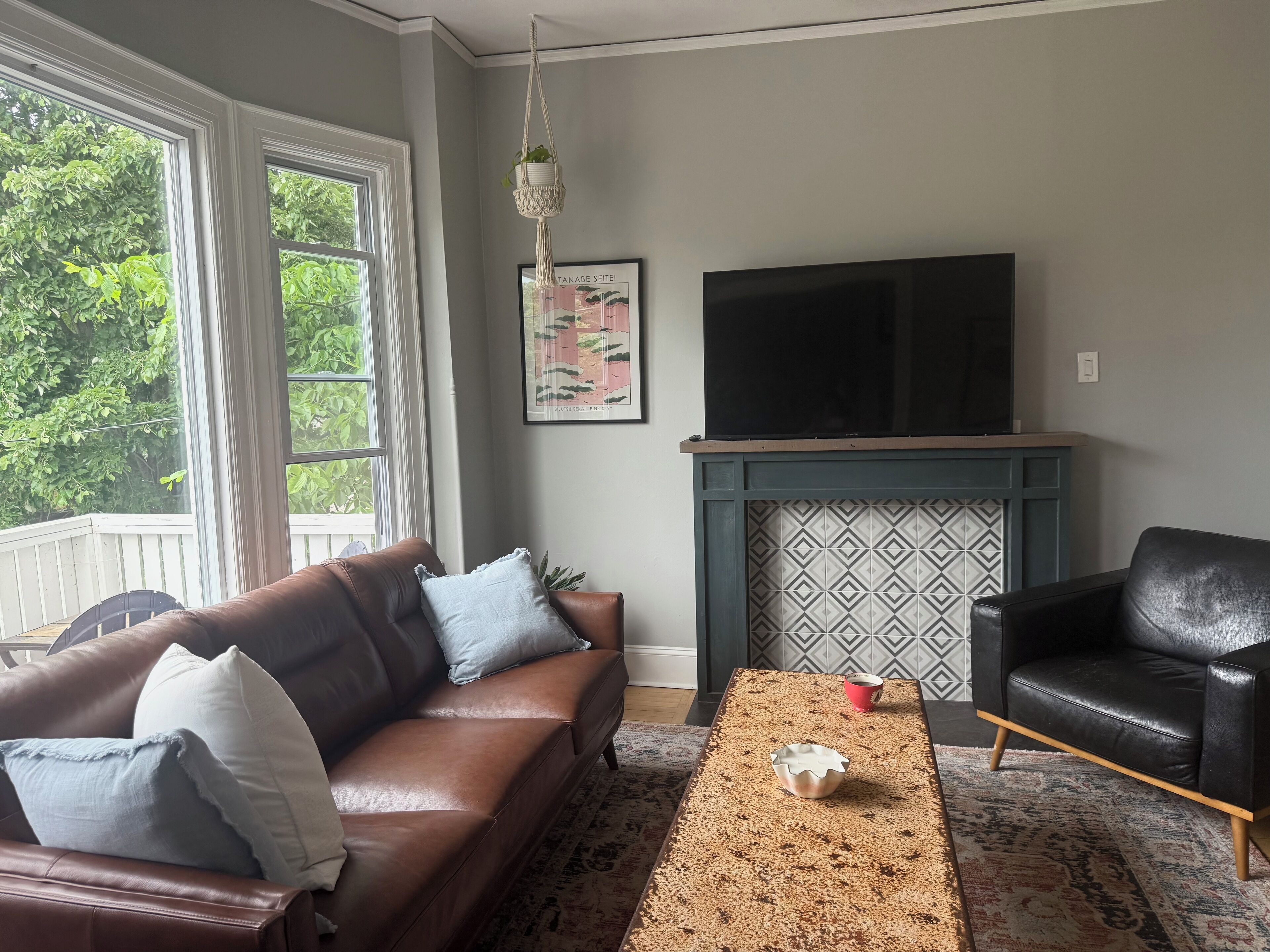 A living room with a brown leather sofa, a black leather armchair, a patterned rug, a fireplace with a decorative mantel, and a large flat-screen TV mounted above the mantel.