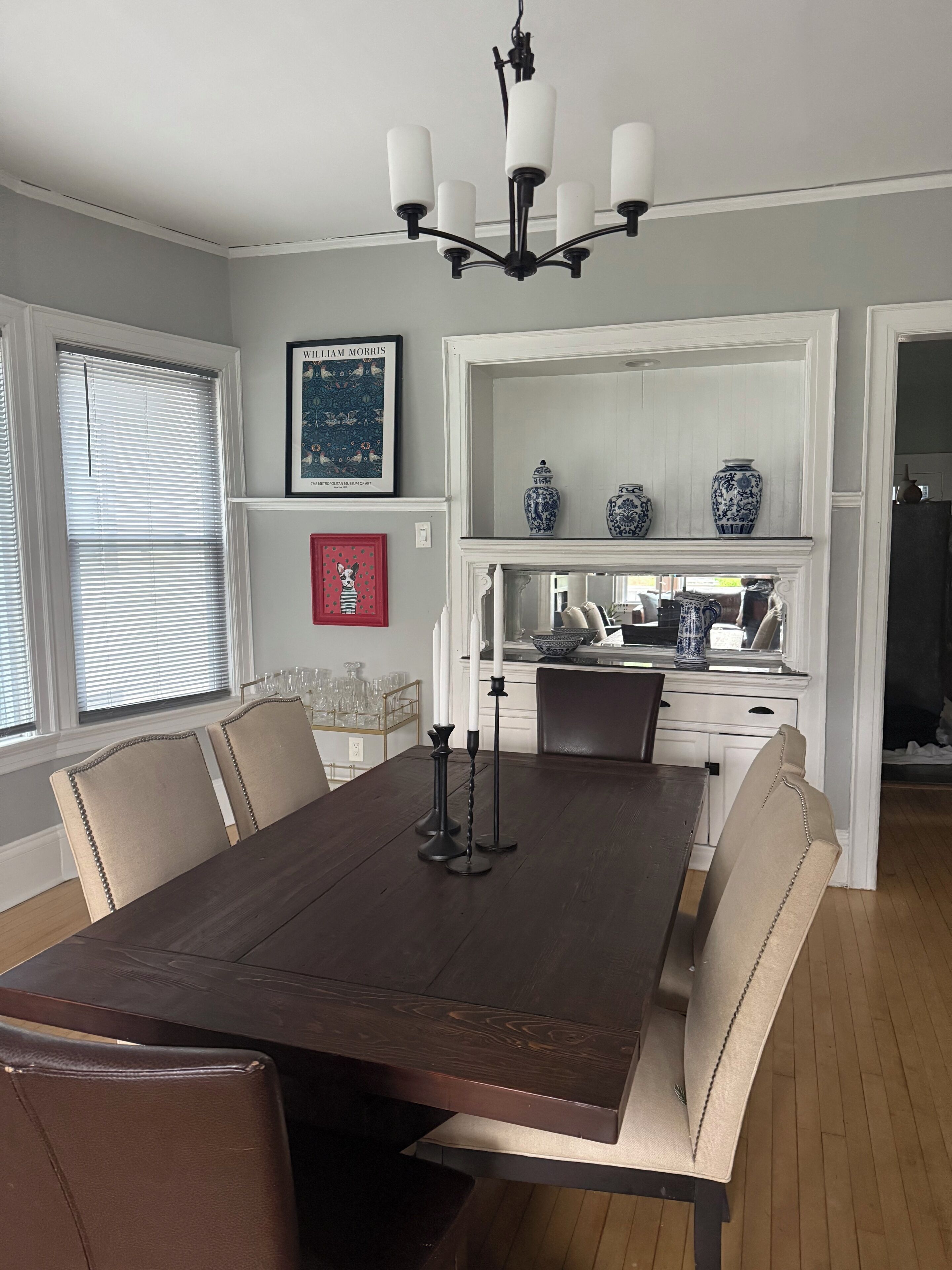 A dining room with a dark wooden table, beige upholstered chairs, a chandelier, and a kitchen area visible through an open doorway.