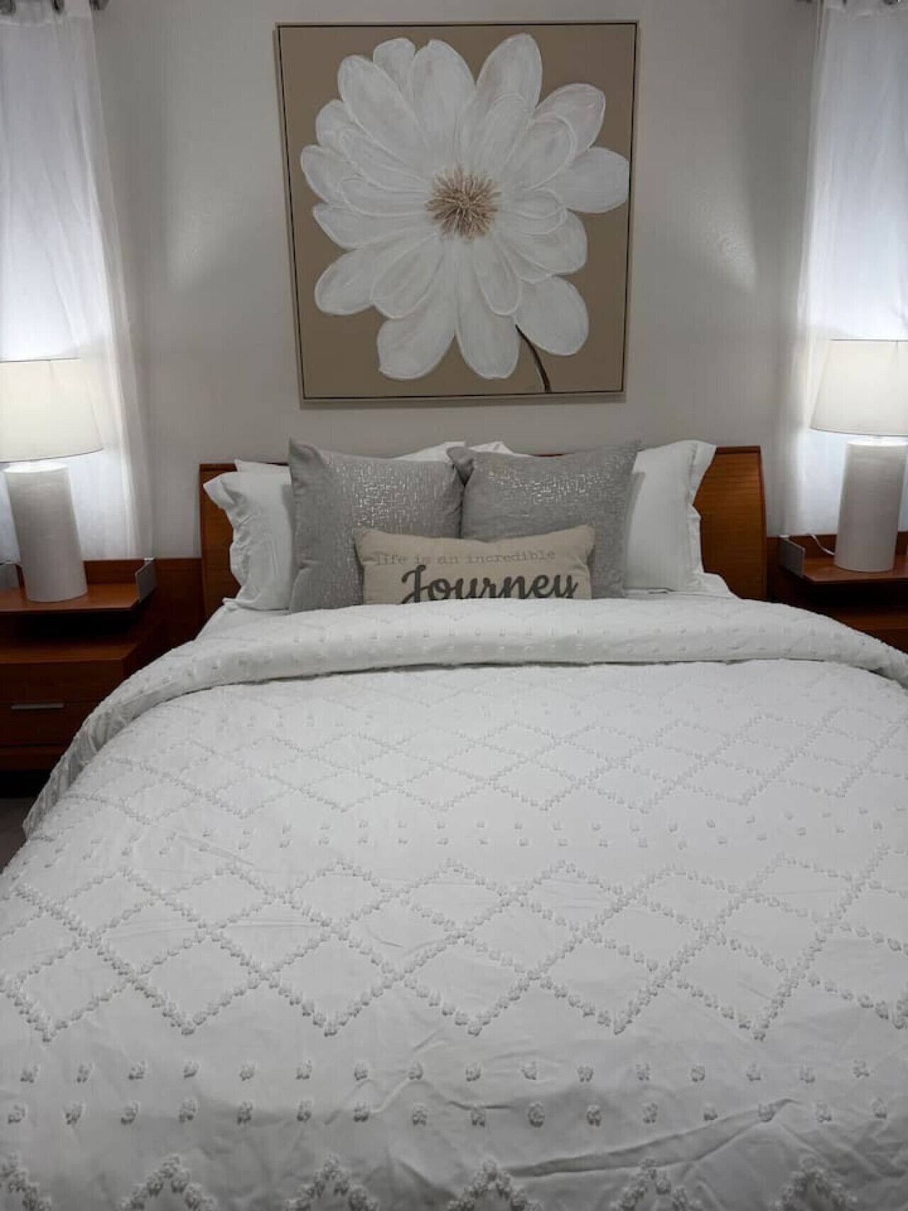 A bedroom with a large bed, a pillow with the word 'Journey,' and a wall-mounted flower painting.