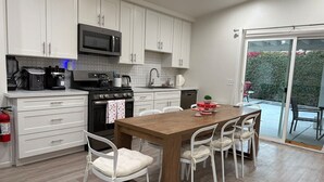 A modern kitchen with white cabinets, a wooden dining table, and a view of the outdoors. - The Coachella Gem with Spa and Fire-pit sleeps 14 (Coachella)