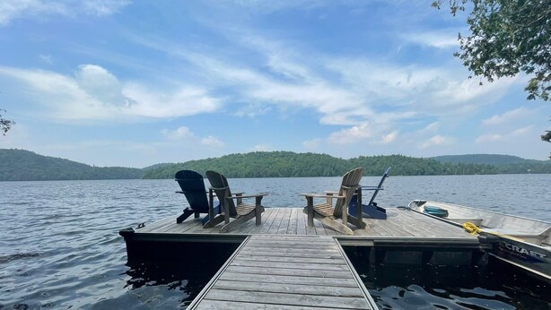 A wooden dock extending into calm water with two Adirondack chairs and a boat docked at the end. - Lakefront 5-Bedroom Chalet in Mont-Blanc - Monthly Rental (Laurentians) (Mont-Blanc)