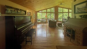 A spacious living room with a wooden ceiling, large windows, a piano, and a fireplace. - Lakefront 5-Bedroom Chalet in Mont-Blanc - Monthly Rental (Laurentians) (Mont-Blanc)