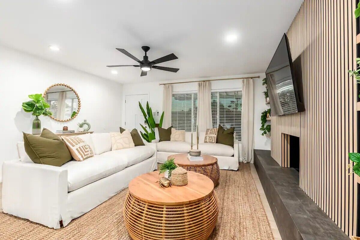 A modern living room with a white sofa, two round wooden coffee tables, a ceiling fan, a flat-screen TV mounted on the wall, and a large window with blinds.