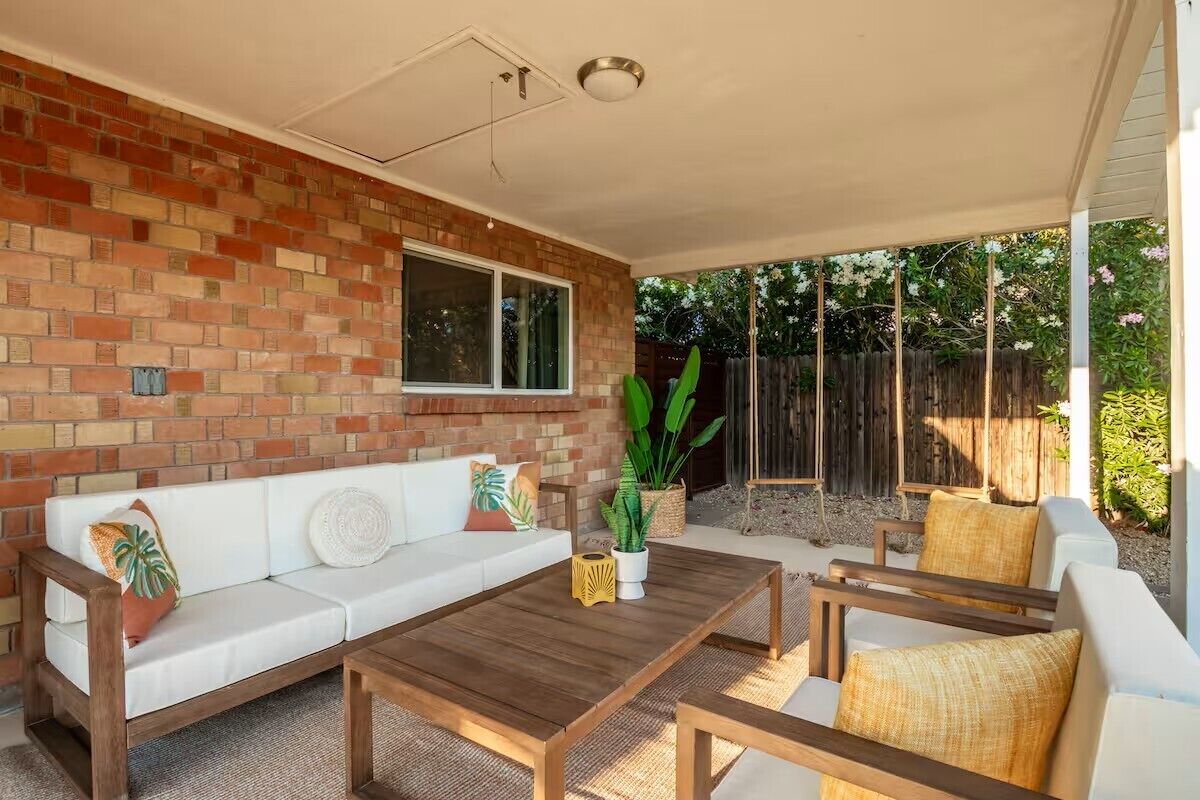 A covered patio area with a brick wall, a white sofa, wooden furniture, and a large plant.