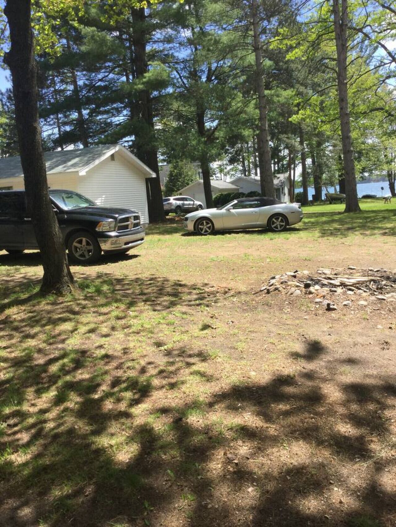 A campsite with two cars parked near a house, surrounded by trees and a lake in the background.