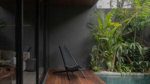 Une terrasse en bois avec un fauteuil de relaxation noir, une porte vitrée et une piscine entourée de plantes tropicales.