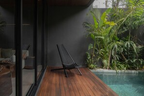 A wooden deck with a black lounge chair, a glass door, and a pool surrounded by tropical plants. - The Akasha Berawa Luxury Villas (Tibubeneng)