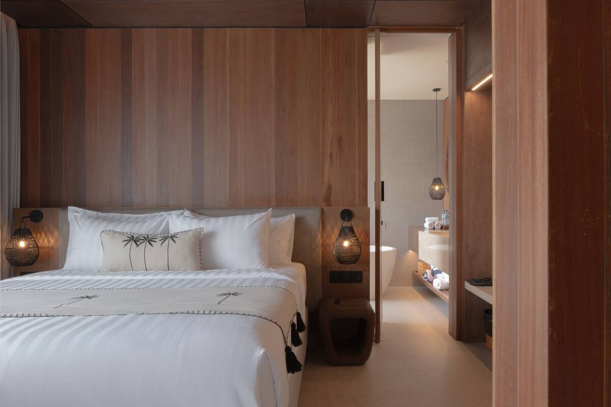 A modern hotel room with a wooden headboard, a bed with white linens, a nightstand with a lamp, and a small seating area with a stool.