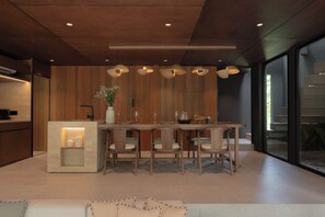 A modern kitchen with wooden cabinetry, a central island with seating, pendant lighting, and a view through large windows. - The Akasha Berawa Luxury Villas (Tibubeneng)