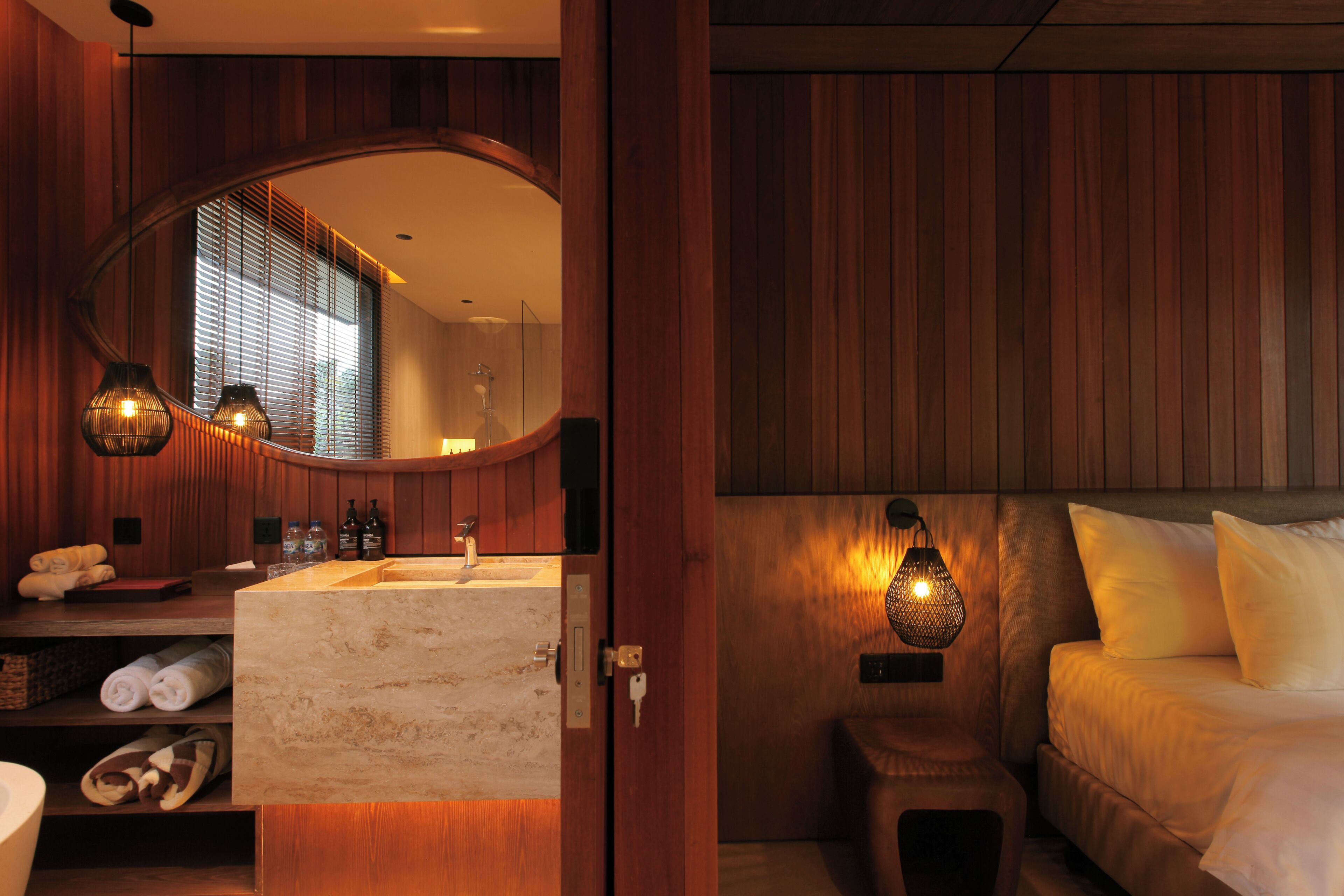 Habitación de hotel con detalles de madera, tocador de baño de mármol, cama con ropa de cama blanca y dos lámparas colgantes.