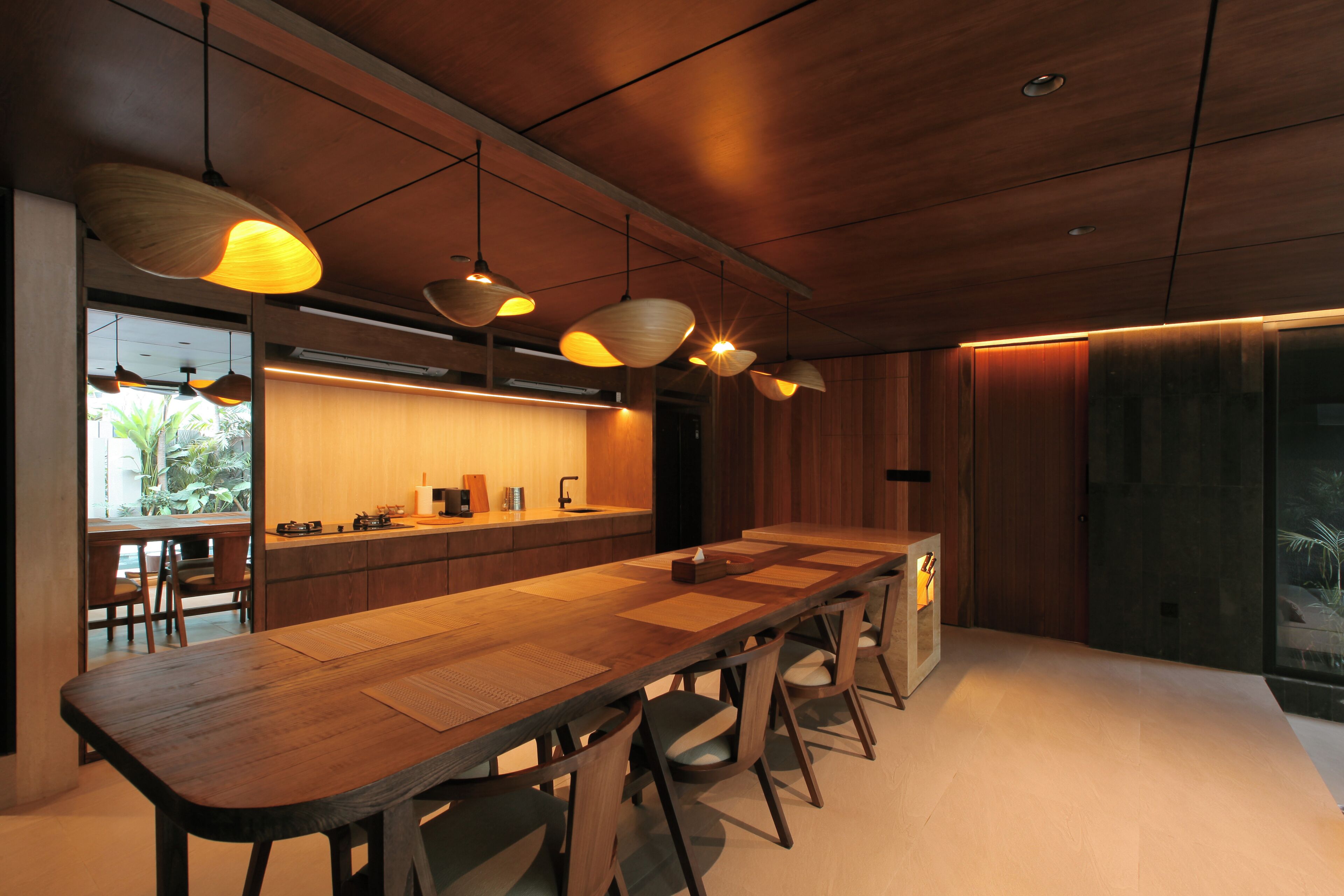 Une cuisine moderne avec une longue table en bois, des luminaires suspendus et une vue sur l’extérieur.