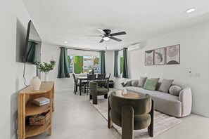 Ein modernes Wohnzimmer mit einem grauen Sofa, einem runden Couchtisch und einem dunkelgrünen Sessel. Es gibt einen Essbereich mit Tisch und Stühlen, und einen Deckenventilator darüber.