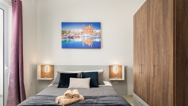 Un letto rifatto con un copriletto grigio scuro e blu, due comodini con lampade a parete, un armadio in legno e un dipinto con una veduta cittadina sopra il letto.