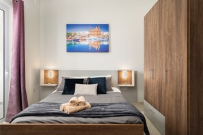 Cama bem arrumada com edredom cinza escuro e azul, duas mesas de cabeceira com luminárias fixadas na parede, guarda-roupa de madeira e uma pintura de paisagem urbana sobre a cama.