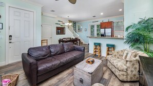 A living room with a brown leather sofa, a wooden coffee table, and a patterned armchair. A ceiling fan and a kitchen area are visible in the background. - OCEAN VIEW ANGLERS REEF - POOL, BEACH. Walk or bike to local food + bars (Islamorada)