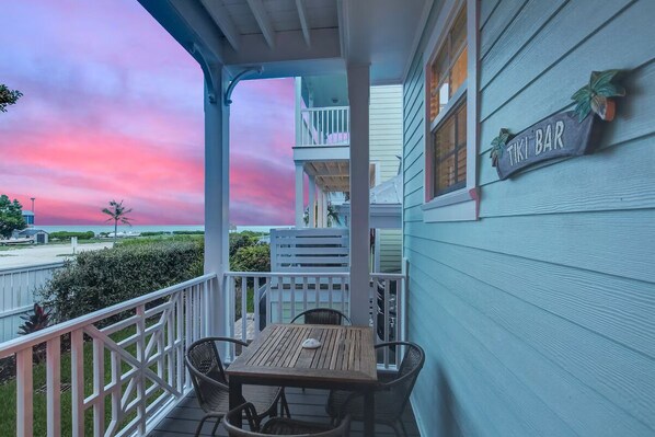 Une véranda abritée avec une table et des chaises, une enseigne indiquant « TIKI BAR », et une vue sur l’océan au coucher du soleil.
