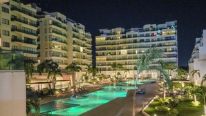 Um hotel com vários andares, piscina, palmeiras e varandas bem iluminadas.