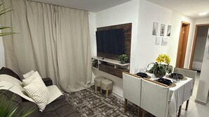 Un salon moderne comprenant un canapé, une table à manger pour quatre personnes, un téléviseur à écran plat fixé au mur et un petit coin cuisine.