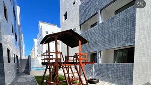 Un immeuble d’appartements moderne doté d’une allée pavée, d’une aire de jeux avec des balançoires et d’un petit espace gazonné.