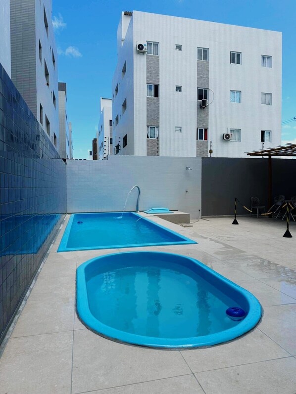 Un área de piscina en la azotea con dos piscinas rectangulares y una circular, rodeada por un edificio de apartamentos moderno.