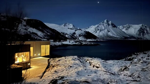En modern byggnad med en fasad av glas och en varm, inbjudande belysning, belägen i ett snöigt landskap med berg i bakgrunden.