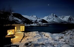 A modern building with a glass facade and warm interior lighting, situated in a snowy landscape with mountains in the background. - Nice home in Bjarkøy with sauna (Bjarkøy)