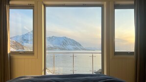 Ett rum med en säng och stora fönster med utsikt över ett snötäckt berg och en sjö eller ett annat vattendrag.