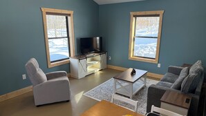 En stue med TV, sofa og lænestol, store vinduer med udsigt til træer, og et lille bord med en vase blomster.