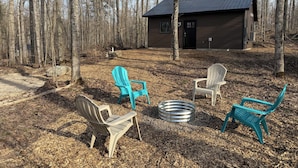 En hytte med metal tag, omgivet af skov. Fire Adirondack stole og en metal pejseplads.