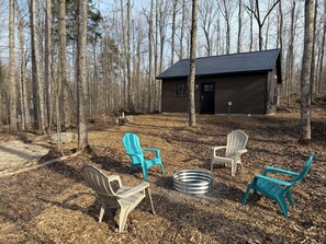 A cabin with a metal roof, surrounded by a forest. There are four Adirondack chairs and a metal fire pit.