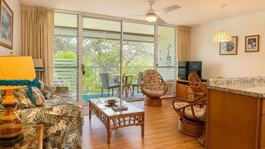 A living room with a sofa, a table, a TV, wicker chairs, and a balcony with a view. - Relaxing Kihei Getaway - Walk to Beaches, Dining & Shops (Kihei)