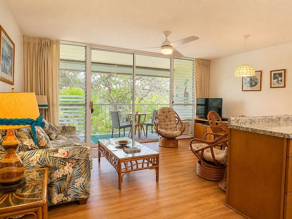 A living room with a sofa, a table, a TV, wicker chairs, and a balcony with a view. - Relaxing Kihei Getaway - Walk to Beaches, Dining & Shops (Kihei)