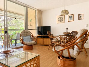 A living room with wicker furniture, a glass coffee table, a TV on a wooden cabinet, and a balcony with a view of greenery. - Soothing 1-bedroom condo with AC in lovely Kihei (Kihei)