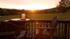 Una terraza de madera con una mesa y sillas, un farol y vistas a un campo verde al atardecer.