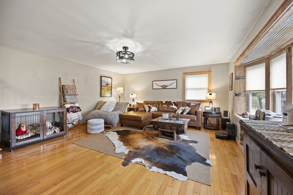 A spacious living room with a wooden floor, a leather sofa, a coffee table, and a large fur rug. There is a dog crate and a lamp on a side table.