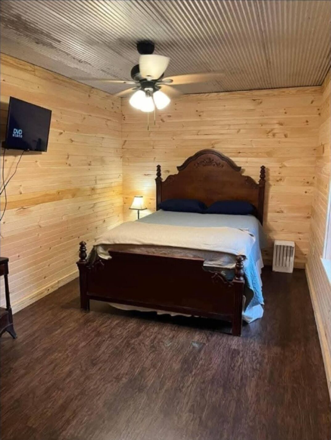 Une pièce aux murs lambrissés, comprenant un lit simple, un ventilateur de plafond, un téléviseur fixé au mur et une petite table.