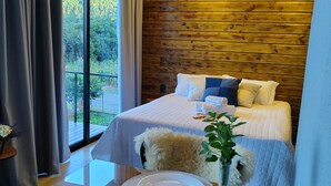 A modern hotel room with a large bed, wooden panel walls, a dining table with plates, and a view of greenery outside. - Cabin in the countryside of Nova Petrópolis, Serra Gaúcha - Chacara Villagio das Pedras (Nova Petrópolis)