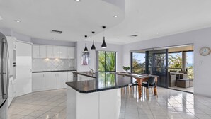 Une cuisine moderne avec des meubles blancs, un îlot central, des luminaires suspendus et une vue sur un balcon aménagé avec du mobilier d’extérieur.