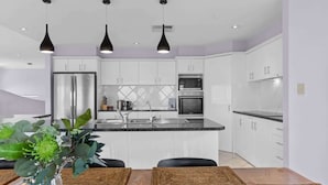 Une cuisine moderne avec des armoires blanches, un réfrigérateur en acier inoxydable, un four à micro-ondes et un four encastré. On y trouve trois luminaires suspendus, un évier et une table à manger avec des sets de table.