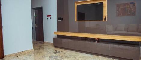 A lobby with a marble floor, a wooden bench, a wall-mounted TV, and a door with a red light.