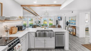 A modern kitchen with white cabinetry, a large island with a sink, a stove, and a dishwasher. There are various kitchen utensils and appliances on the countertops. - Steps to Sand: Ground-Floor Condo with Patio, Private Beach Access & Pool! ☀️ (Satellite Beach)