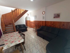 A cozy living room with a wooden staircase, two sofas, a dining table with floral tablecloth, and a small wooden chair. - Holiday home with air conditioning, private beach section and jetty, large garden (Balatonmáriafürdő)