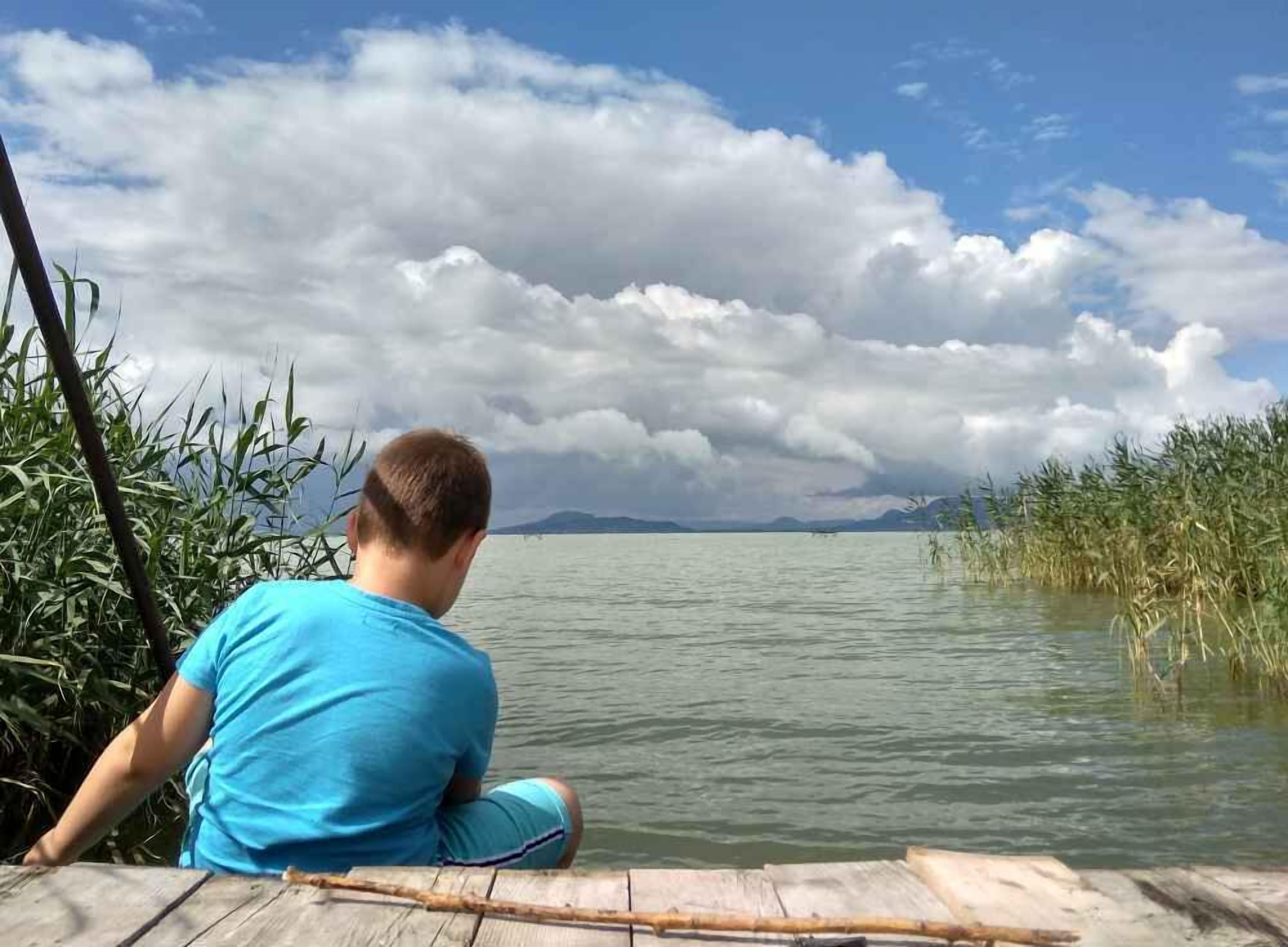 En person sidder på en træbro ved en stille sø med siv langs bredden, under en delvist overskyet himmel.
