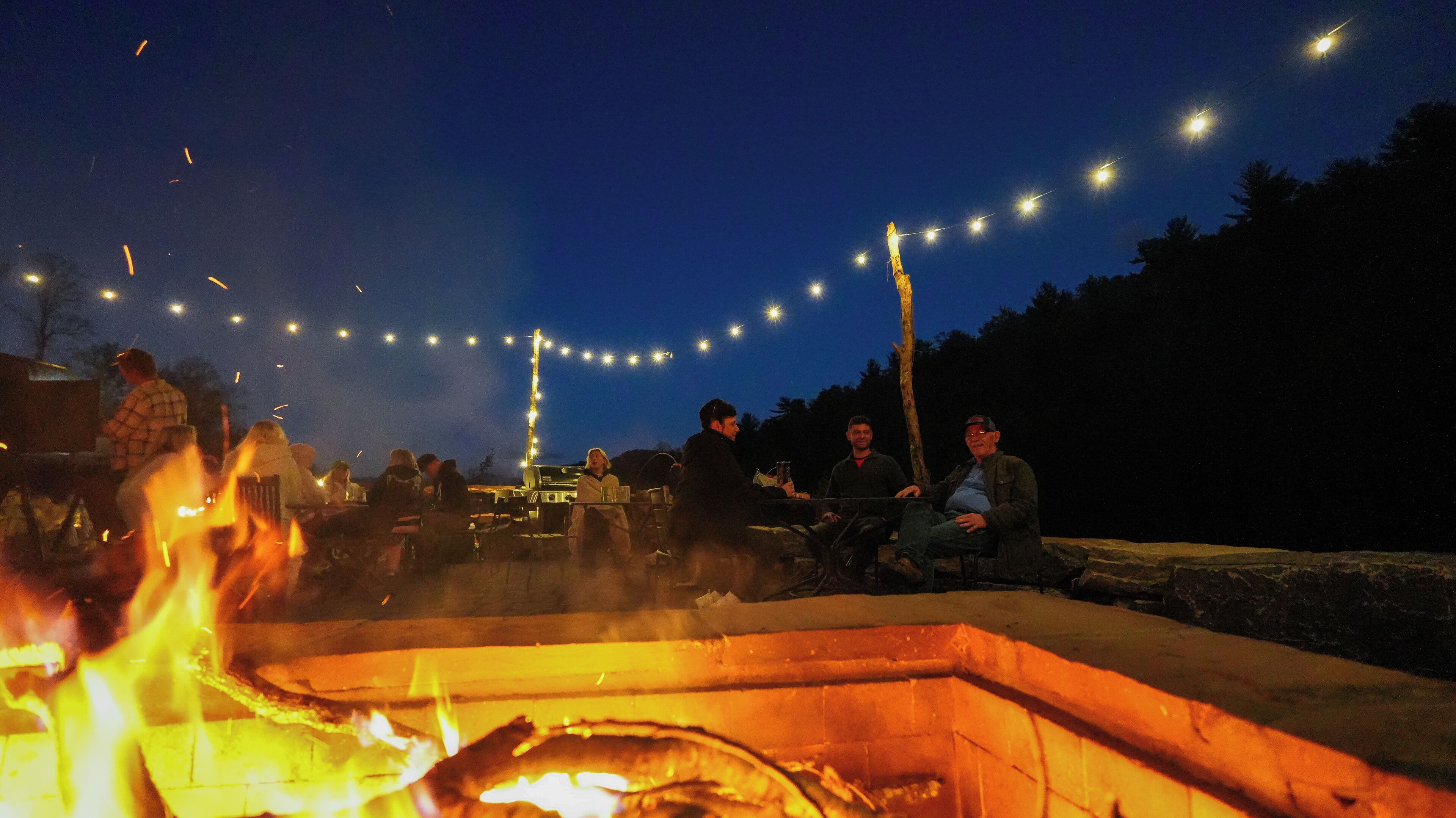 A group of people sitting around a campfire at night with string lights overhead.