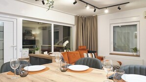 A dining area with a wooden table set for two, two wine glasses, and a plate. There is a window with curtains, a plant on the windowsill, and a view of the kitchen in the background. - House in Međimurje for couples with jacuzzi, sauna, terrace, pool, and barbecue (Dragoslavec)